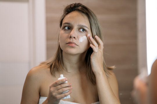 Woman applying moisturizing cream to her face while looking in a bathroom mirror. Skincare routine, facial care, and morning beauty ritual.