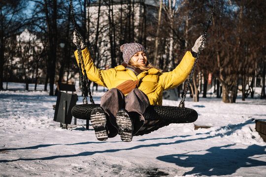 Young woman in yellow jacket smiling and enjoying a swing on a winter day in a snowy park, surrounded by cold bright weather.