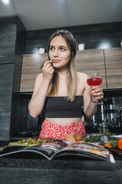 Woman sipping an alcoholic cocktail or rose wine while reading a cookbook and preparing ingredients at the kitchen counter. Evening culinary hobby, relaxed lifestyle, and home cooking.