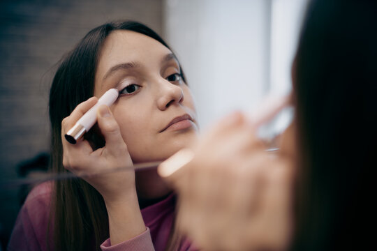 Young woman applying eyeshadow with a makeup pencil while looking in the mirror. Concept of daily beauty routine, cosmetics, skincare, self-care, and personal grooming at home.
