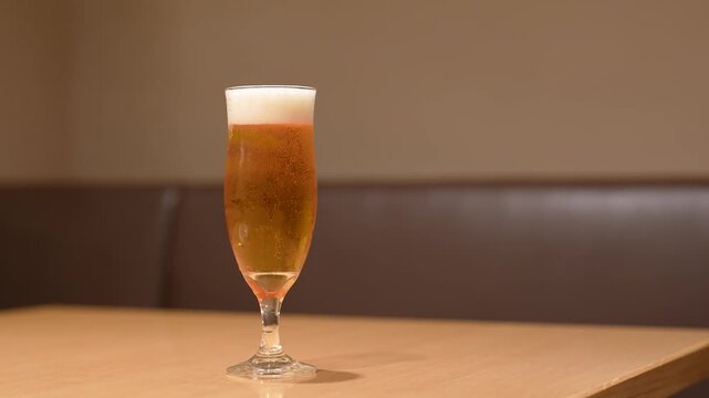 A glass of amber lager beer in tulip glass is placed on wooden table, with foam on top and bubbles floating up. Drink object footage, close-up with selective focus.