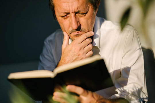 Man absorbed in reading a book outdoors on a sunny balcony, surrounded by plants, leisure and relaxation concept with natural light.