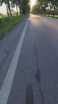 Vertical cycling POV footage along a quiet rural road lined with trees and green fields, with warm golden hour sunlight glowing at the end of the road.