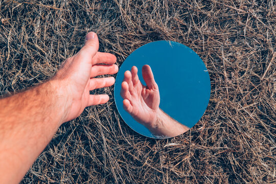 Top view of a hand reaching toward a round mirror lying on dry grass, reflecting the hand and blue sky, concept of reflection, self-image and perspective