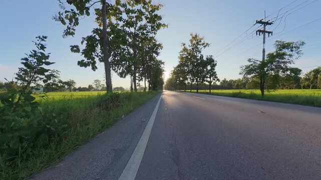 Rural asphalt road running through green fields at sunset, with roadside trees, overhead power lines and a motorcycle in the distance under a clear blue sky.