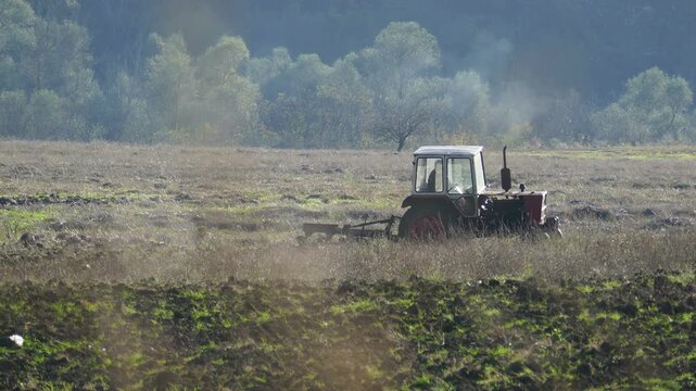 Distant shot of an old Soviet-era vintage tractor maneuvering in a field with an unrecognizable driver, historic Eastern Bloc agriculture machinery working on a farm plantation in Bulgaria