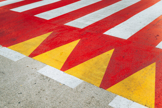 A high-angle close-up shot of vibrant red, white, and yellow geometric road markings painted on asphalt, featuring zebra crosswalk stripes and triangular yield markings.