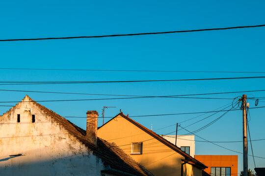 Residential house roofs and utility wire infrastructure against a bright blue sky at sunset. Concept of suburban living, neighborhood power grid, telecommunication, and housing development.