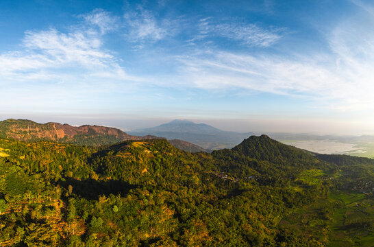 Aerial view of Mount Telomoyo with the distant Mount Ungaran, featuring lush forested hills, rugged cliff edges, and a vast landscape in Getasan, Central Java, Indonesia.