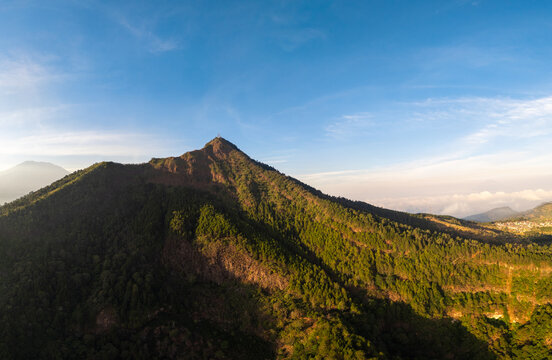 Aerial view of Mount Telomoyo with lush green forested slopes under a clear blue sky during golden hour in Getasan, Central Java, Indonesia.