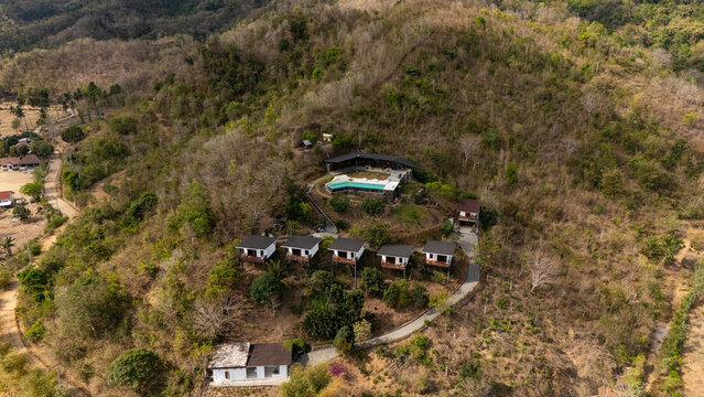 Aerial view of a hillside resort with modern villas, a central pool, and a winding driveway, set amidst dry tropical vegetation in Praya Barat, West Nusa Tenggara, Indonesia.