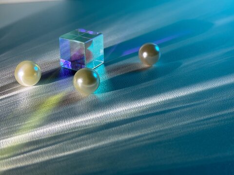 Transparent glass cube and three imitation pearls illuminated by blue and purple light, creating colorful reflections, refractions and long dramatic shadows on a textured surface.