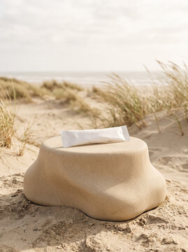 Sculptural sandy-beige pedestal, blank white paper stick sachet. Outdoor coastal setting, blurred dune grasses and sandy landscape. Mockup.