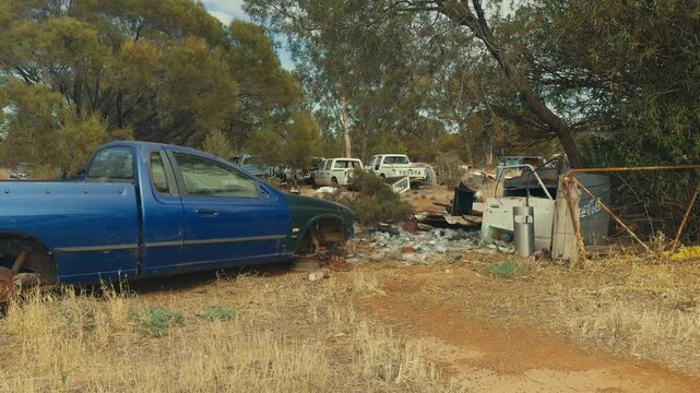 Abandoned blue ute with missing wheels next to piles of discarded bottles and a rusty gate in an outback wrecking yard among eucalyptus trees