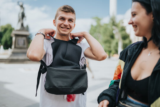 A smiling teenage boy puts on a backpack while talking to a female friend. They relax and chat together outdoors in an urban square on a sunny day.