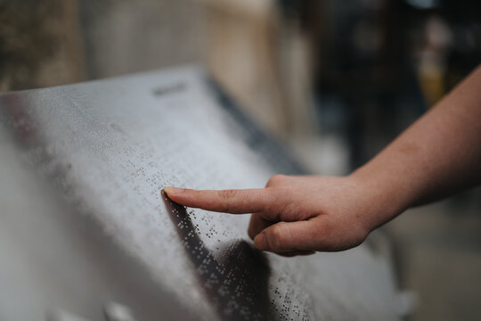 A person uses a fingertip to read raised braille dots on a metal plaque. The close-up shows tactile reading and accessible signage for people with visual impairments.