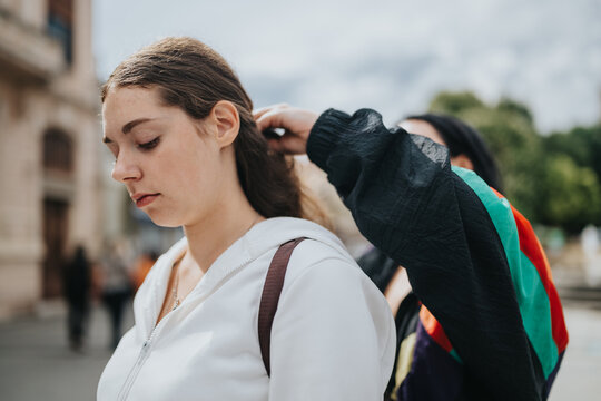 A young woman stands as a friend adjusts her hair outdoors in an urban setting. The pair share a quiet, casual moment on a city street.