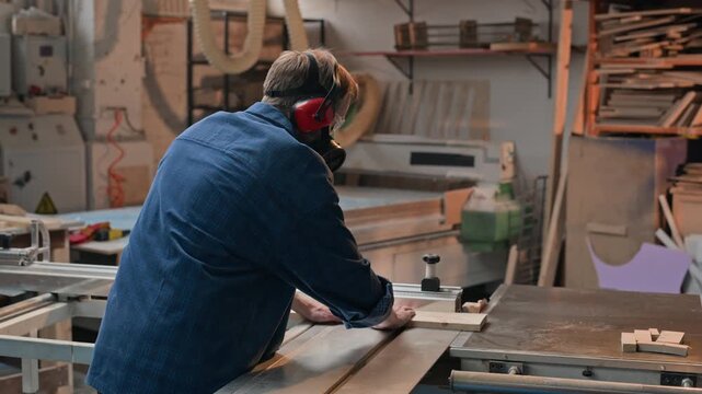 Caucasian middle aged craftsman operating sliding table saw in woodworking shop from rear view. Clip suitable for workplace safety training and craft industry marketing