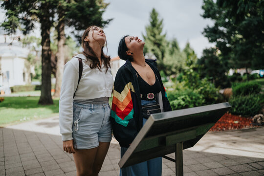 Two young women stand by an outdoor information board, both looking upward. They appear to be sightseeing together in a leafy park setting.