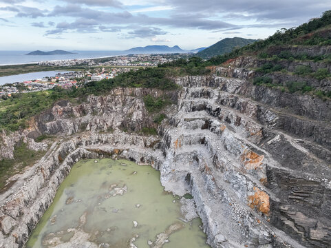 Aerial view of Pedreira Rio Tavares, a large stone quarry with terraced rock faces and a pool of water at the base, situated near the coastline in Florianopolis, Santa Catarina, Brazil.