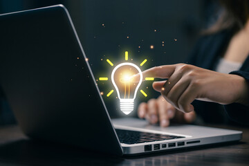 Business professional touching a glowing light bulb innovation icon above a laptop, symbolizing creative thinking, new ideas, problem solving, digital transformation and modern business development. © saranyoo