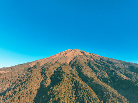Aerial view of the majestic Mount Sumbing peak with lush green forested slopes and rugged terrain under a clear blue sky in Kecamatan Kaliangkrik, Jawa Tengah, Indonesia.