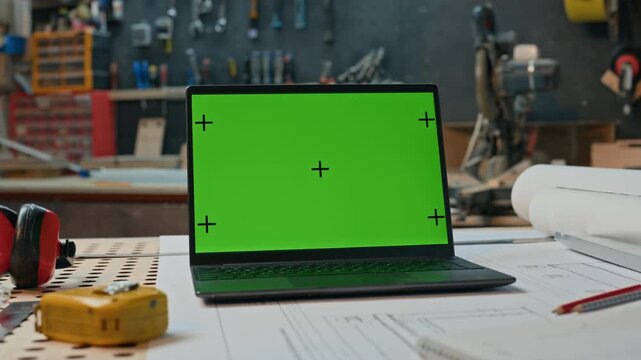 Laptop displaying green screen mockup with tracking markers resting on woodworking workshop table near blueprints. Ideal for displaying architectural design software interface