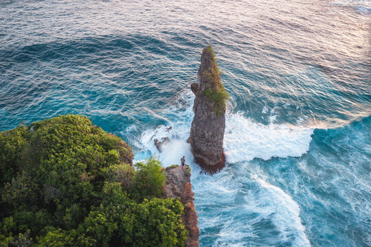 Aerial view of a person standing on a cliff edge overlooking a tall sea stack surrounded by turquoise ocean waves and lush green vegetation in Nusa Penida, Bali, Indonesia.