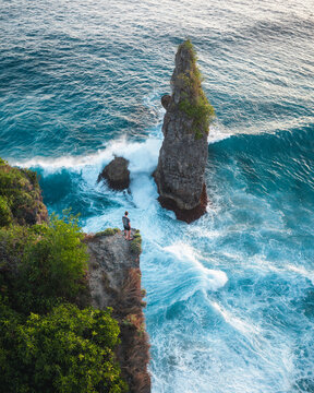 Aerial view of a lone person standing on a cliff edge overlooking a dramatic sea stack surrounded by turquoise ocean waves crashing against the rocks in Nusa Penida, Bali, Indonesia.