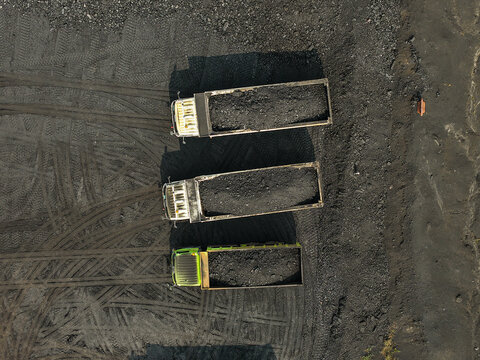 Aerial view of three dump trucks loaded with coal parked on a dark mining site with visible tire tracks in the ground in Tenggarong, East Kalimantan, Indonesia.