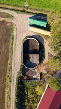 Aerial view of an oval-shaped slurry lagoon surrounded by agricultural fields, a dirt road, covered hay bales, and a building with a red roof in Milan, Lombardy, Italy.