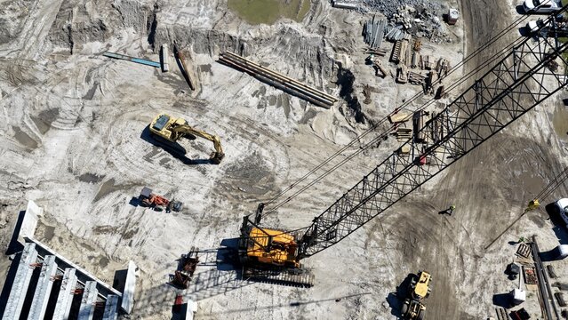 Aerial view of a construction site featuring a large yellow crawler crane, an excavator, and heavy machinery on sandy ground in St. Johns County, Florida, United States.