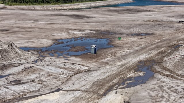 Aerial view of a solitary portable toilet standing in a large, muddy construction site with tire tracks and puddles in St. Johns County, Florida, United States.