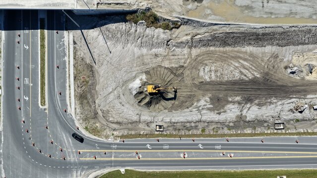 Aerial view of an excavator working on a construction site with piles of dirt, surrounded by a paved road with traffic cones and a vehicle in St. Johns County, Florida, United States.