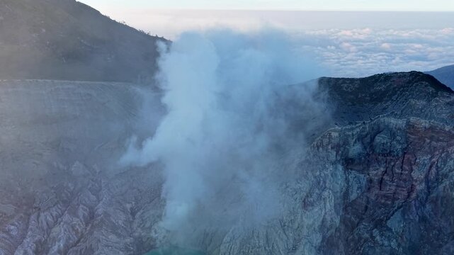 Voluminous smoke billows from volcanic crater. Rugged cliffs frame steaming, turquoise waters at Mount Ijen
