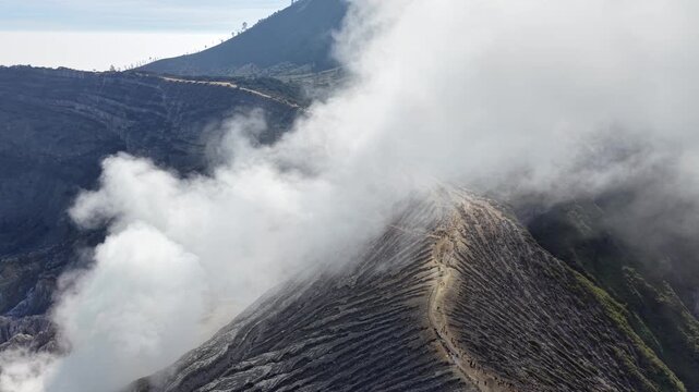 Volcanic smoke curls from rugged, dark terrain. Layered ridges frame the smoky crater edge at Mount Ijen
