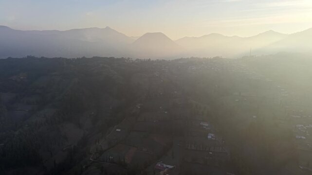 Aerial drone view of misty sunrise over volcanic valleys and rural farmland in Wonotoro Sukapura.