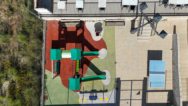 Aerial view of a colorful playground with slides and climbing equipment on a paved terrace next to a restaurant patio, Murla, Valencian Community, Spain.