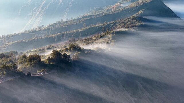 Misty dawn wraps mountain ridges in soft, flowing haze. Low sun bleaches sky at Cemoro Lawang