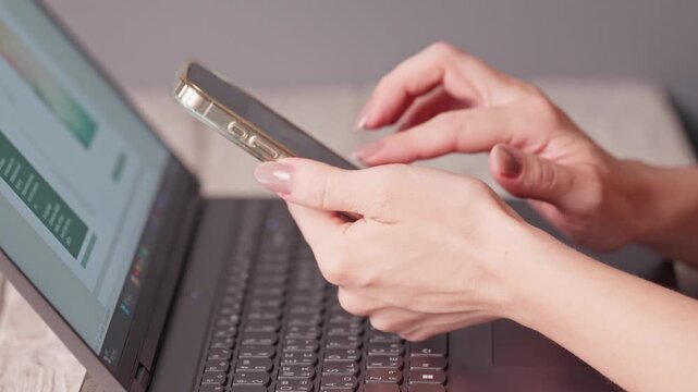 Close-up of female hands holding a smartphone and scrolling social media feed next to an open laptop. Concept of multitasking, procrastination, mobile technology, freelance work and digital habit