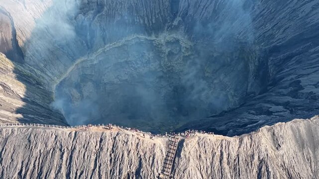 Drone aerial over Mount Bromo crater showing steaming fumaroles, rugged rim textures and visitors on narrow pathway.