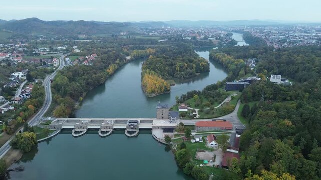 Aerial view of the Drava River and Ototok hydroelectric power plant in Maribor, Slovenia, featuring a concrete dam, a tree-covered island with golden autumn foliage, and the city of Maribor in the bac