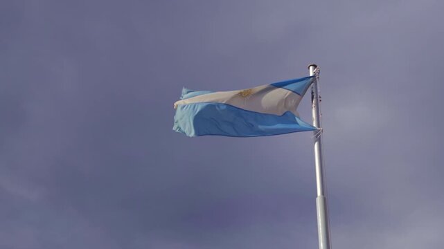 Low angle static shot of the blue and white Argentine flag fluttering in strong wind on a tall white flagpole under overcast sky.