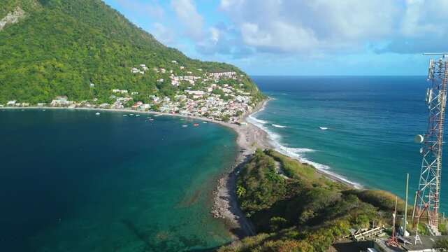Aerial view as camera flies forward along narrow sandbar toward Scotts Head village with calm bay on left and ocean waves on right in Dominica