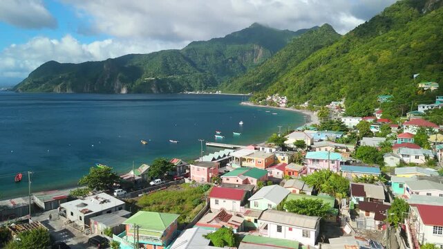 Aerial view as camera moves slowly forward over colorful houses and fishing boats of Scotts Head village along blue bay beneath lush green mountains in Dominica