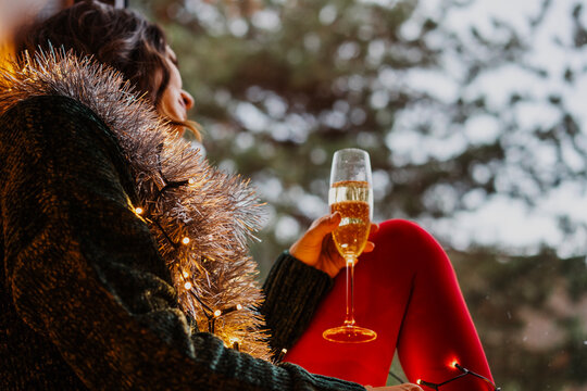 Side view of a beautiful young woman in a green sweater and red tights sitting on a windowsill with a champagne glass.