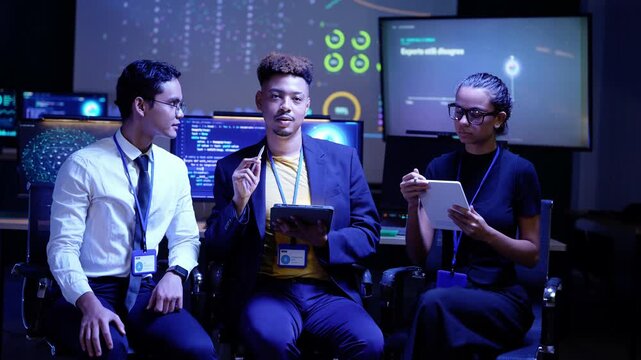 Black developer in a navy blazer talks with a stylus in hand while an Asian colleague in a tie listens and a Latina woman writes on a tablet, seated in a dark AI lab