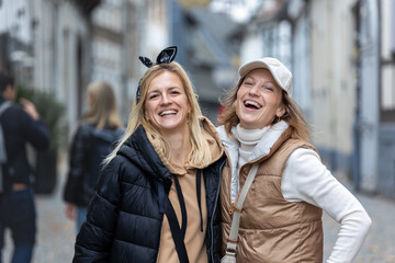 Two happy women laugh together while posing on cobblestone street, wearing jackets during autumn...