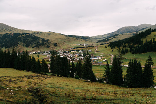 Serene mountain village nestled between rolling green hills. Dark evergreen trees line the valley floor, framing the small settlement. Clouds drift over distant peaks. Ak-Suu, Jyrgalan, Kyrgyzstan
