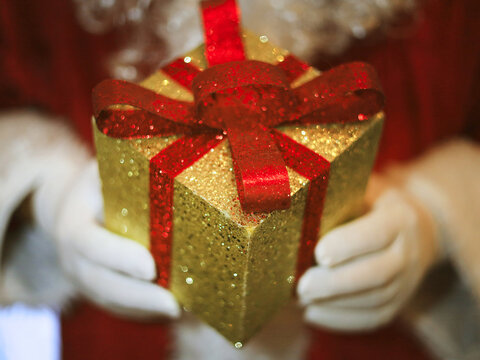 Gold gift box with a red glitter bow rests in Santa's white gloves, linking festive Christmas packaging to seasonal gift shopping and the holiday tradition of choosing wrapped presents for exchange.